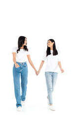 full length view of happy interracial women in jeans holding hands while walking on white.