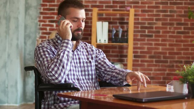 Person with disabilities is sitting on wheelchair at table with laptop and talking on phone. Concept of solving business issues remotely