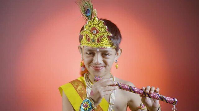 Smiling Indian little kid in lord Krishna costume playing flute by looking at camera - concept of hindu tradition, celebration of shri krishna Janmashtami festival