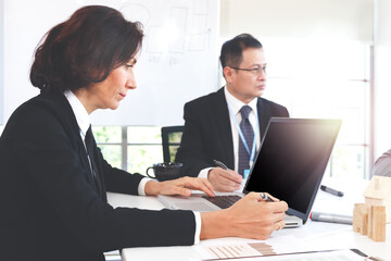 Middle aged business woman uses laptop computer during brainstorming discussion at meeting table in office with senior Asian businessman background, variety multi-ethnic team colleagues work together