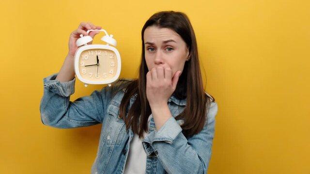 Portrait of impatient young caucasian female 20s biting her nails and holding clock in hand, deadline, wearing blue denim jacket, posing isolated over pastel yellow color background wall in studio