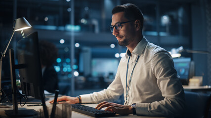 In Big Corporate Office at Night: Portrait of Confident Manager in White Shirt Using Computer, Businesspeople and Experts Working Around Him, Analysing Statistics, Commerce Data, Marketing Plans.