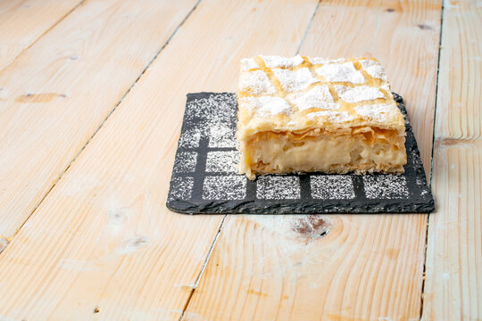 cremeschnitte or cremsnit cake on a square black stone tile texture with sprinkled sugar powder and a white washed wood boards table