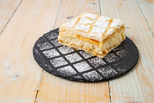 cremeschnitte or cremsnit cake on a square black stone tile texture with sprinkled sugar powder and a round black stone tile, side view