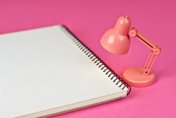 Notepad on a pink background. Toy table lamp near the notebook. Copy space and free space for text near the lamp. The concept of roochy space and creative ideas.