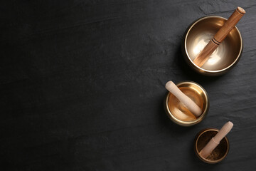 Golden singing bowls and mallets on black table, flat lay. Space for text