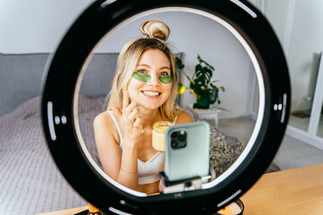 Beauty blogger using ring lamp and making photo or video content for social media with smartphone in bedroom at home. Blond woman portrait through light of ring lamp.