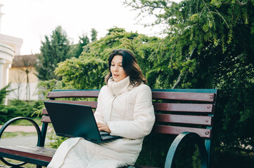 Young woman of Asian appearance searching job with a laptop in an urban park, freelancer. Online communication, online business. The woman blogger works on a laptop computer in the office.