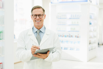 Successful male mature chemist pharmacist druggist in white medical coat looking at camera writing...