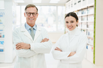 Two caucasian confident successful male and female chemists pharmacists druggists in white medical coats standing with arms crossed looking at camera in pharmacy drugstore