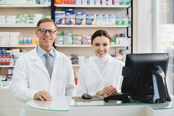Smiling two successful male and female chemists pharmacists druggists in white medical coats standing at the cash point desk looking at camera in pharmacy drugstore