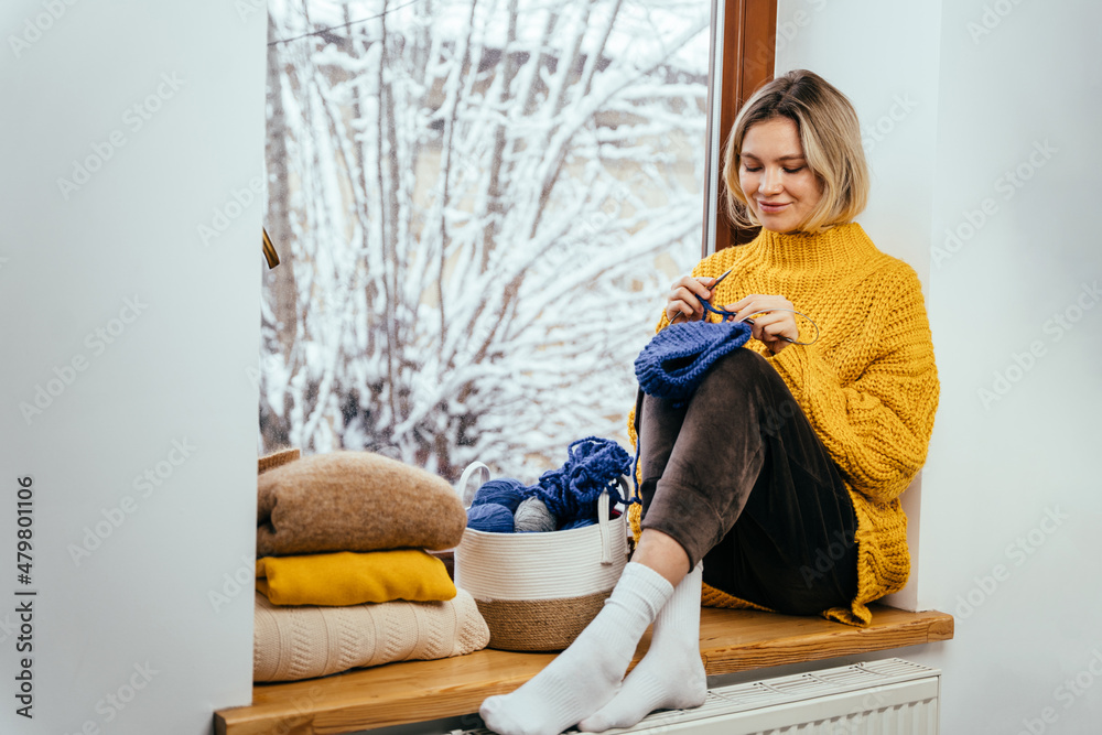 Wall mural Winter portrait of happy young blond woman wearing yellow knitted sweater. Girl holding knitting needles a basket of knitted clothing sitting on window sill.