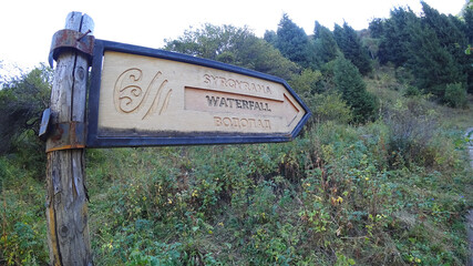 A post with a wooden pointer to the waterfall. Coniferous green forest, bushes and grass grow. We are following the path in the right direction. A sign with information in three languages. Kazakhstan