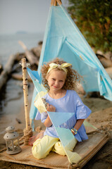 Smiling girl with curly hair sitting on the shore with whigwam on background. © Yuliya Gorbunova