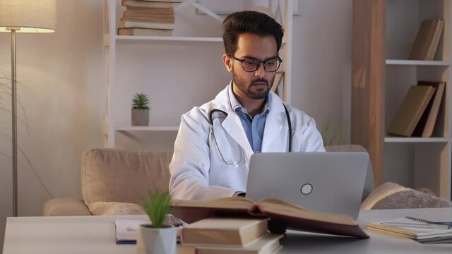 Male doctor. Doctoral thesis. Educational research. Smart man typing scrolling laptop sitting desk with books light room interior.