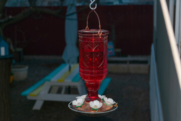 red glass hummingbird feeder hanging on the side of a house during the late evening