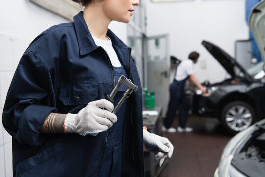 Cropped view of mechanic in uniform and gloves holding tool in car service.