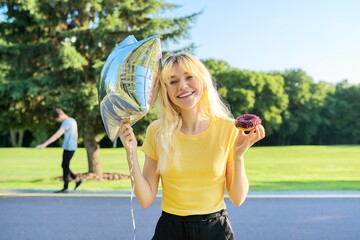 Portrait of beautiful teenage blonde with cake donut and silver balloon in park