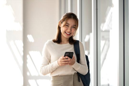 Pretty smiling happy young Asian student standing outside the classroom hand holding smartphone.  Looking at camera.
