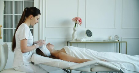 Curing skin problems. Female cosmetologist looking at client's face and giving a facial massage. Happy relaxed young woman getting professional facial treatment in spa salon. High quality 4k footage