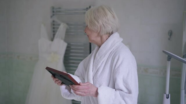 Beautiful senior Caucasian woman with grey hair looking back at wedding dress in bathroom admiring picture in photo frame. Portrait of happy smiling mother on daughter's wedding day indoors