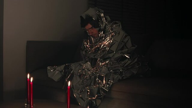 Alone man suffers from electromagnetic hypersensitivity and hides under a metal blanket in dark room with candles. Man is afraid of electrical appliances