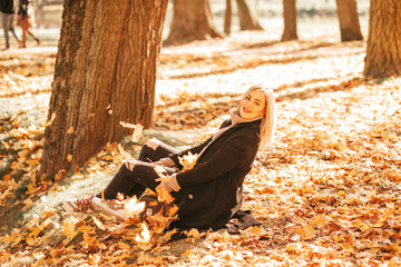 Golden autumn. Positive blonde in black coat and woolen sweater with large collar poses sitting on old suitcase in autumn forest . Fun while hiking in autumn season.