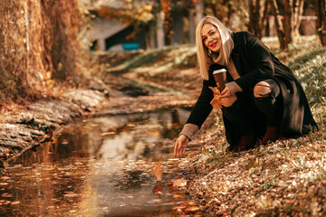 A charming blonde in fashionable clothes, sitting down among bright foliage of city park, stretches her hand to the golden ribbon of which the stream has become. She has coffee in her hands