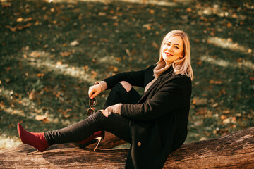 Woman in warm cozy clothes is relaxing sitting in autumn park on fallen tree in rays of golden sun. Beautiful women. Autumn portrait. Recreation and travel. Enjoying nature. Weekend.