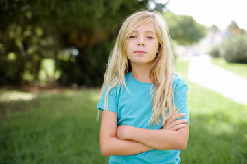 Serious pensive beautiful Caucasian little kid girl wearing blue T-shirt standing outdoors feel...