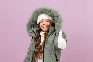 a little girl in a winter jacket and a warm knitted hat gives a thumbs up. a child in winter clothes on an isolated background.