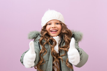 a little girl in a winter jacket and a warm knitted hat gives a thumbs up. a child in winter clothes on an isolated background.