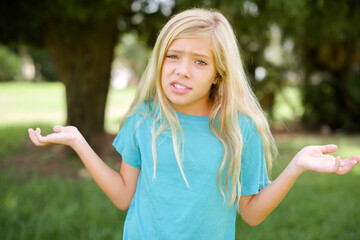 Clueless Caucasian little kid girl wearing blue T-shirt standing outdoors  shrugs shoulders with...