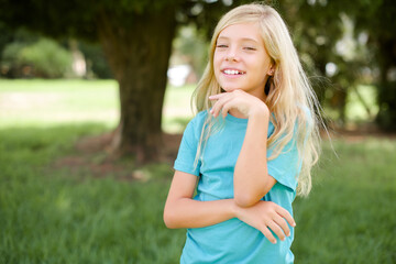 Fototapeta premium Optimistic Caucasian little kid girl wearing blue T-shirt standing outdoors keeps hands partly crossed and hand under chin, looks at camera with pleasure. Happy emotions concept.