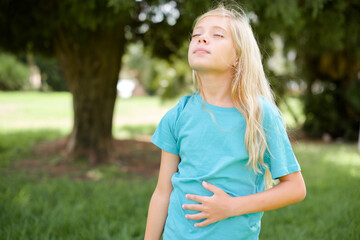 Fototapeta premium Caucasian little kid girl wearing blue T-shirt standing outdoors touches tummy, smiles gently, eating and satisfaction concept.