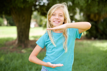 Caucasian little kid girl wearing blue T-shirt standing outdoors gesturing with hands showing big and large size sign, measure symbol. Smiling looking at the camera.