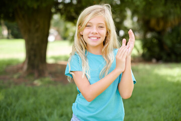 Caucasian little kid girl wearing blue T-shirt standing outdoors clapping and applauding happy and...