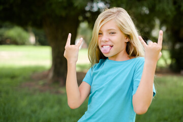 Caucasian little kid girl wearing blue T-shirt standing outdoors  making rock hand gesture and...