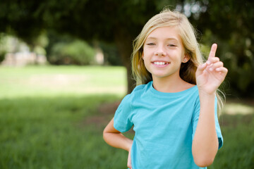 Caucasian little kid girl wearing blue T-shirt standing outdoors holding finger up having idea and posing