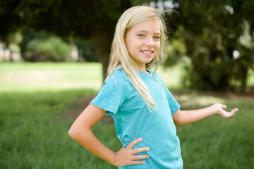 Caucasian little kid girl wearing blue T-shirt standing outdoors feeling happy and cheerful, smiling and welcoming you, inviting you in with a friendly gesture