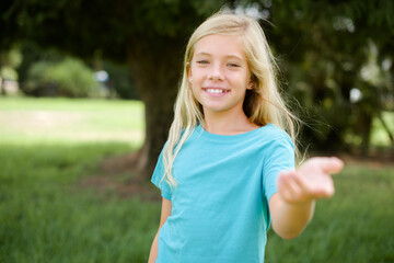 Caucasian little kid girl wearing blue T-shirt standing outdoors smiling cheerful offering palm hand giving assistance and acceptance.