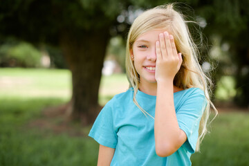 Caucasian little kid girl wearing blue T-shirt standing outdoors covering one eye with her hand, confident smile on face and surprise emotion.