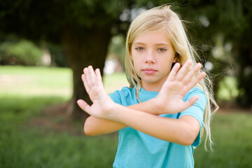 Caucasian little kid girl wearing blue T-shirt standing outdoors has rejection expression crossing arms and palms doing negative sign, angry face.