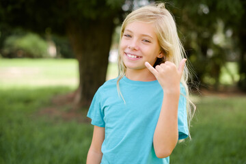 Caucasian little kid girl wearing blue T-shirt standing outdoors smiling doing phone gesture with hand and fingers like talking on the telephone. Communicating concepts.