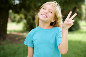 Caucasian little kid girl wearing blue T-shirt standing outdoors smiling with happy face winking at...