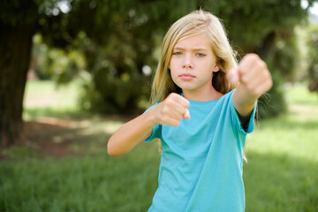 Caucasian little kid girl wearing blue T-shirt standing outdoors Punching fist to fight, aggressive and angry attack, threat and violence