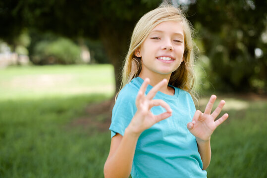 Caucasian little kid girl wearing blue T-shirt standing outdoors showing both hands with fingers in OK sign. Approval or recommending concept