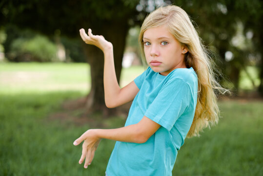 Caucasian little kid girl wearing blue T-shirt standing outdoors pointing aside with both hands showing something strange and saying: I don't know what is this. Advertisement concept.