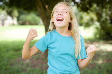 Caucasian little kid girl wearing blue T-shirt standing outdoors celebrating surprised and amazed...