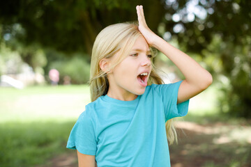 Caucasian little kid girl wearing blue T-shirt standing outdoors surprised with hand on head for...